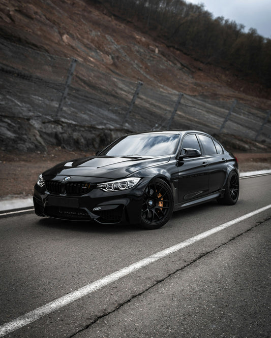 Black F80-generation BMW M3 parked on a paved road, front three-quarter view showing factory ride height